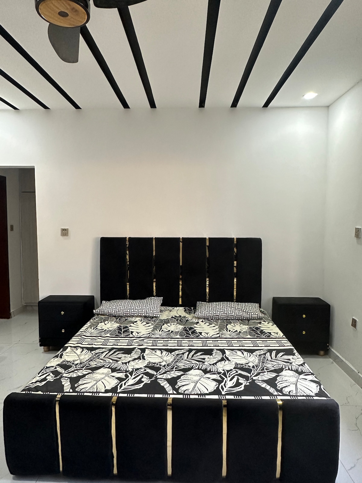 The image features a modern bedroom with a black upholstered bed adorned with a floral patterned blanket. Two matching nightstands are placed on either side of the bed. Ceiling details include dark stripes, and soft lighting highlights the room's simplicity.
