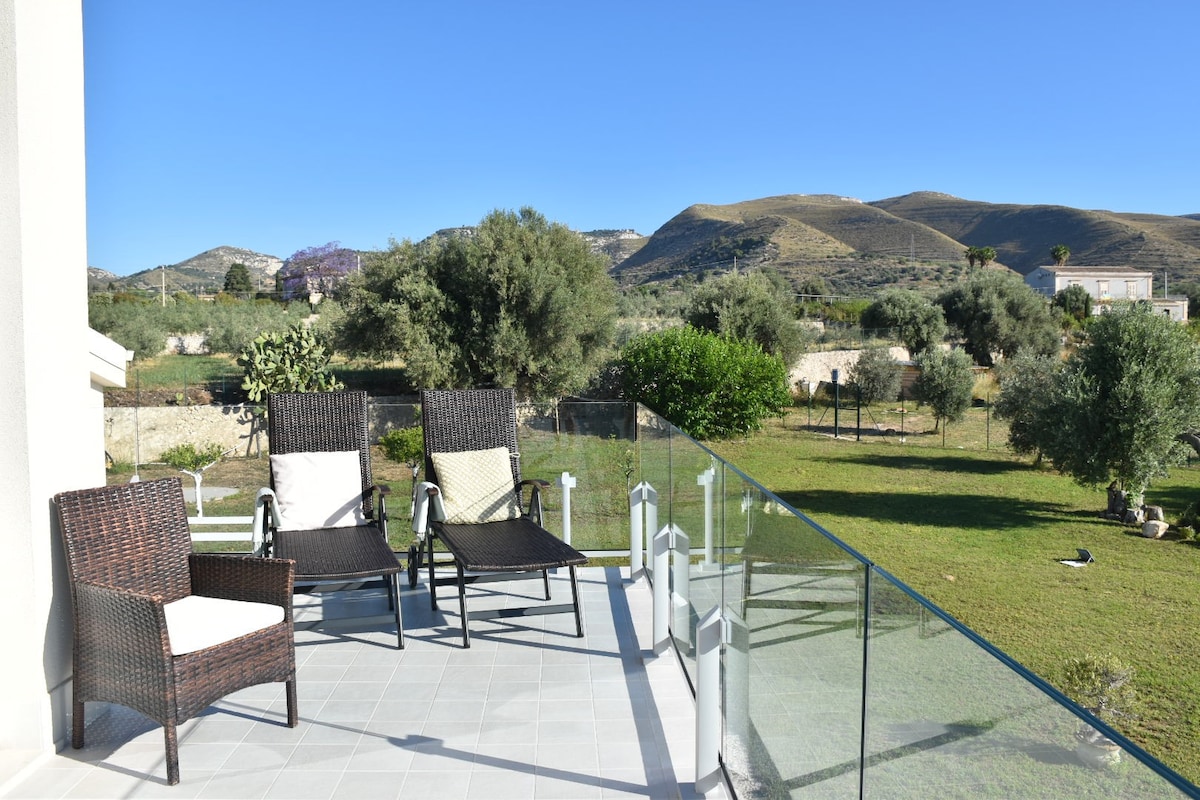 An outdoor terrace features four wicker chairs arranged around a glass balustrade. A lush green lawn and olive trees are visible in the background, with gentle hills rising in the distance under a clear blue sky.