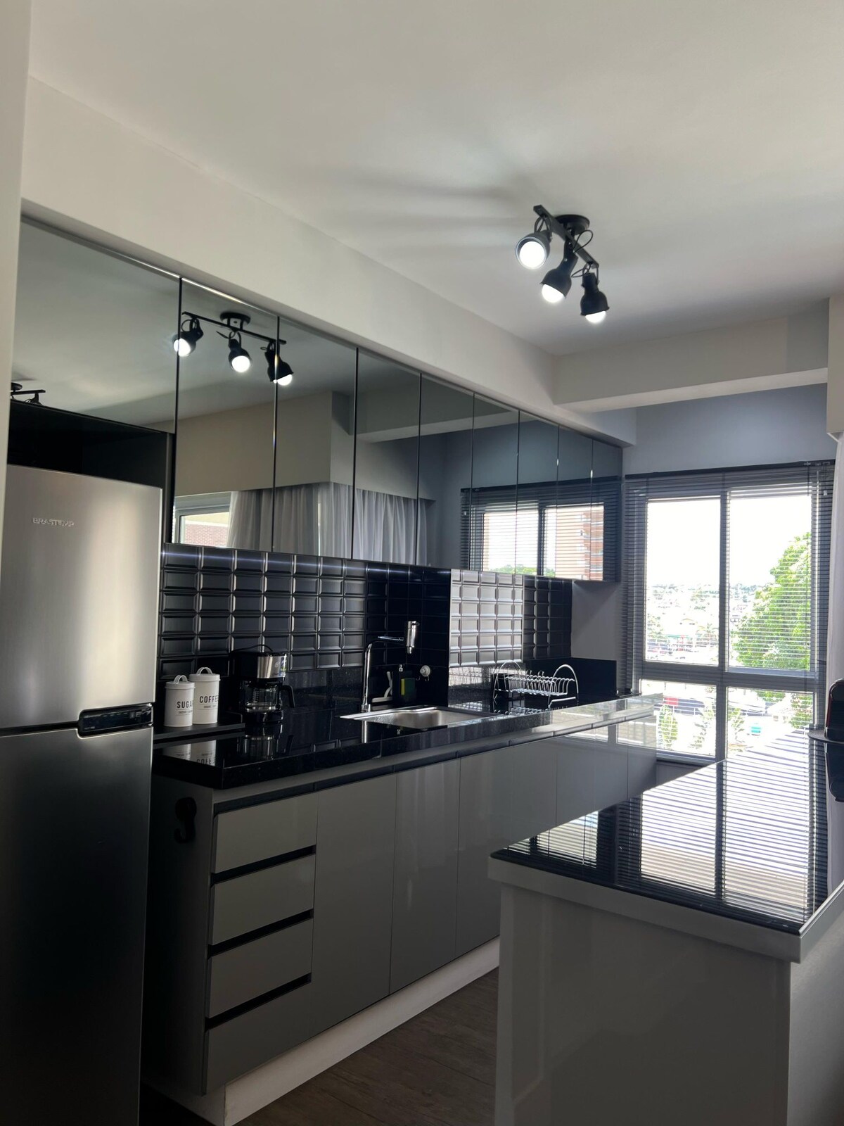 A modern kitchen is showcased with sleek black cabinets and a large island countertop. Stainless steel appliances, including a refrigerator, are neatly arranged, and natural light filters through large windows, illuminating the space. The glossy tiles add a touch of elegance to the overall design.