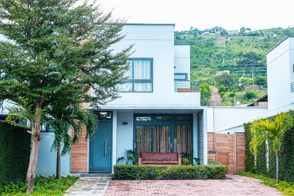 A modern two-story building is framed by lush greenery, with a welcoming front porch featuring a wooden bench. The exterior is complemented by large windows, allowing natural light to fill the interior spaces while offering views of the surrounding hillside.