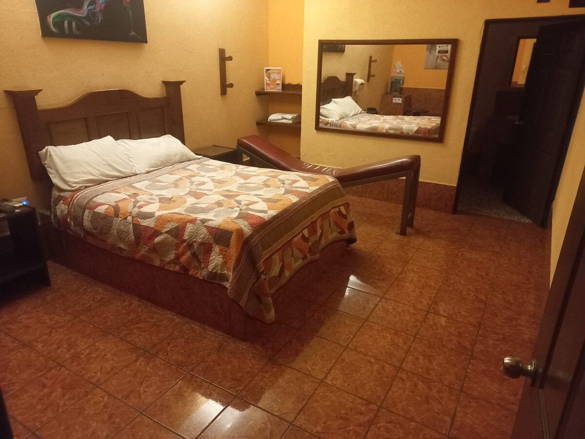 A sleeping area features a spacious bed adorned with a patterned quilt. A mirror reflects the room's warm tones, complemented by tiled flooring. A bench sits at the foot of the bed, and the layout allows for clear movement throughout the space.