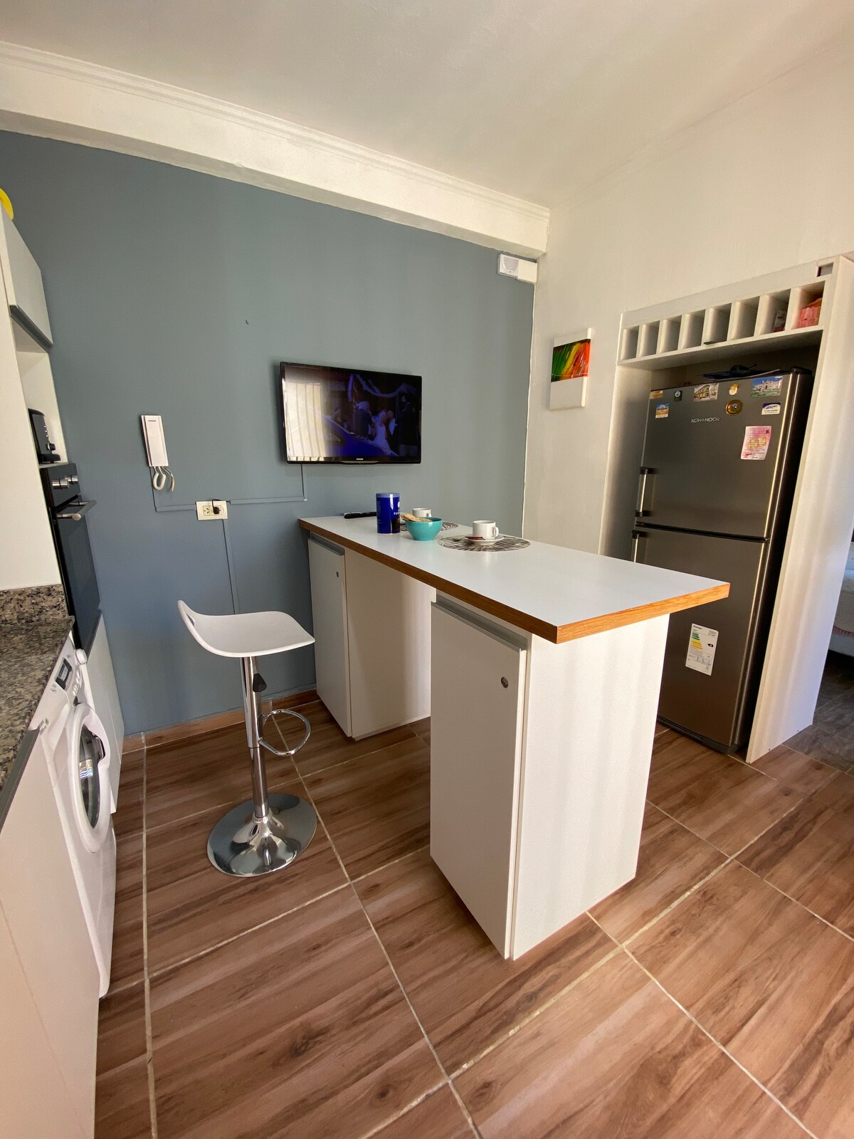 A compact kitchen area is presented with a streamlined bar top and a high stool. A wall-mounted television is located above the bar, while essential appliances, including a refrigerator and washing machine, are visible. Soft, neutral tones create a calm and functional space.