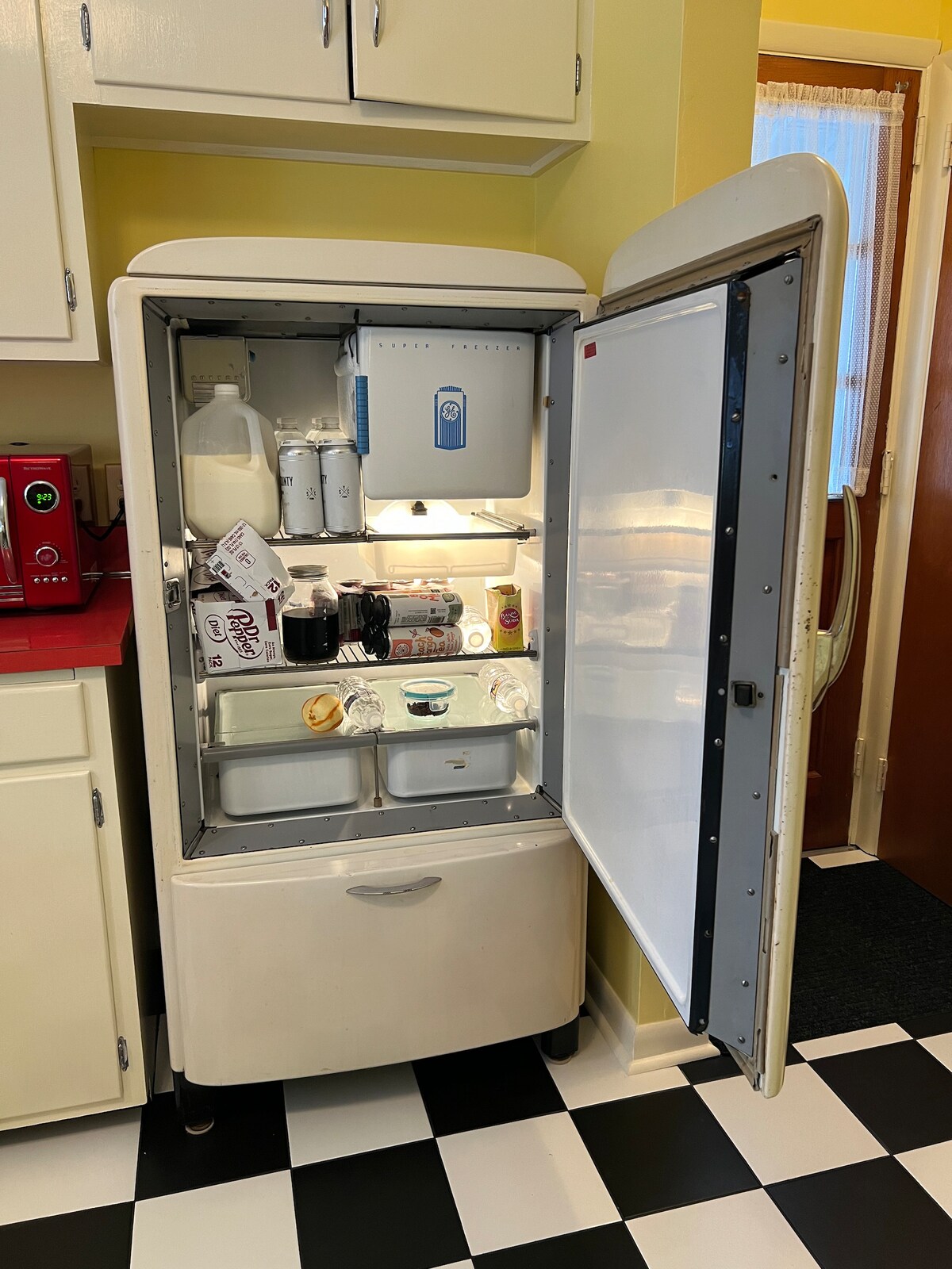 An open vintage refrigerator reveals its contents, showcasing a variety of items including beverages and milk. The interior is well-lit, highlighting the organized shelves within. The refrigerator is framed by classic cabinetry and set against a cheerful yellow wall. A tiled floor adds to the nostalgic ambiance.