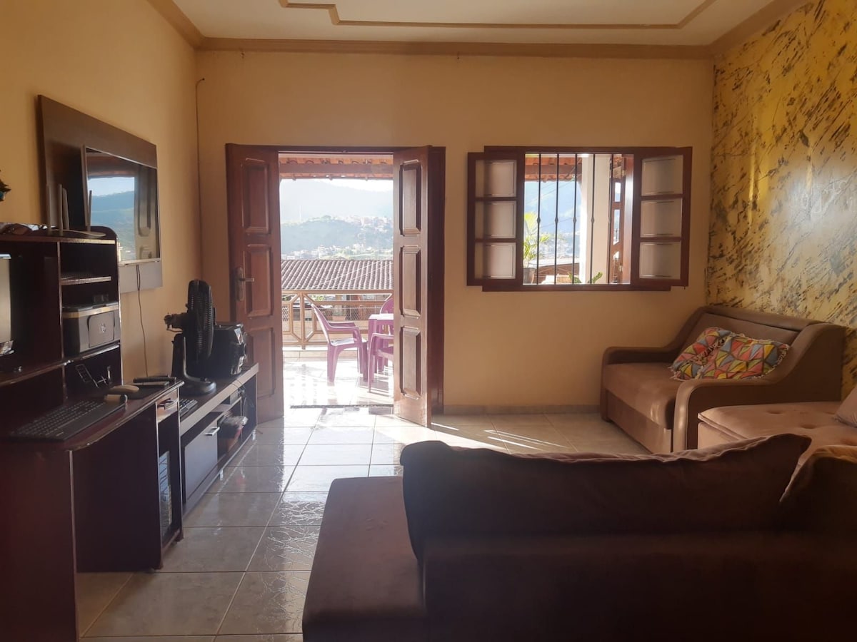 A living area features a comfortable sofa and a small desk with a computer setup. Natural light enters through open double doors, leading to a balcony with outdoor seating. A wall adorned with a colorful map adds visual interest to the space.