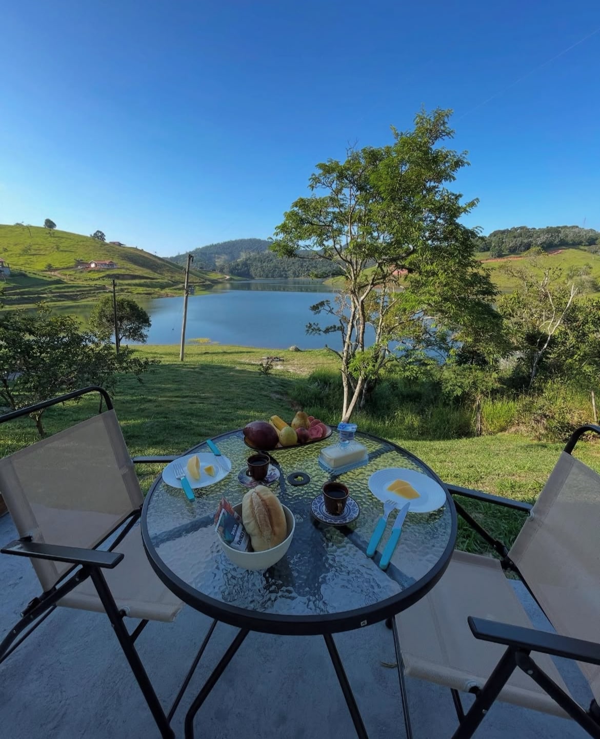A small round glass table is set for breakfast, featuring fruit, bread, and coffee. Two chairs are positioned around the table, with a serene lake and rolling green hills visible in the background under a clear blue sky.