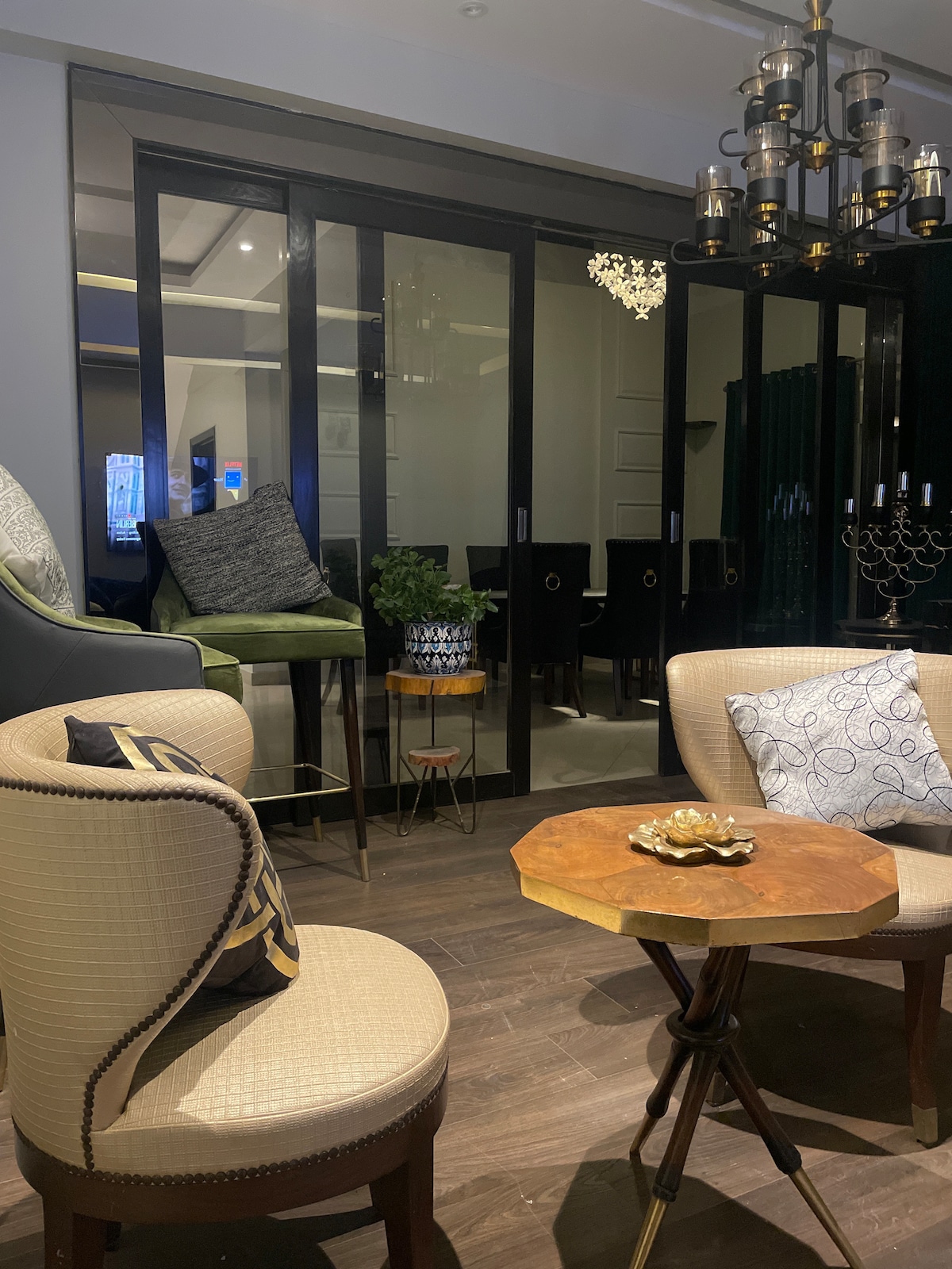 A stylish lounge area features two upholstered chairs and a small wooden table with decorative elements. A chandelier hangs above, and large glass doors provide a view into a well-lit dining space, creating an inviting setting for relaxation.