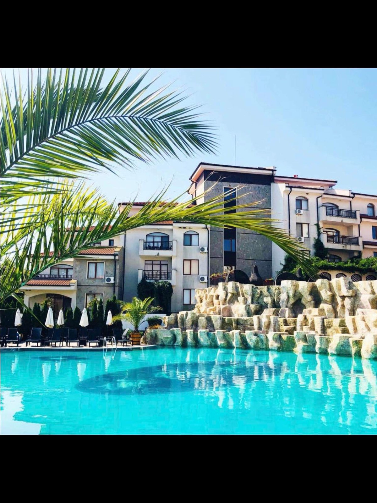 The image displays a large swimming pool with vibrant turquoise water, surrounded by tropical palm leaves. In the background, a multi-storied building is visible, characterized by a modern design and multiple balconies. A rocky feature encircles the pool area, enhancing the resort-like ambiance.