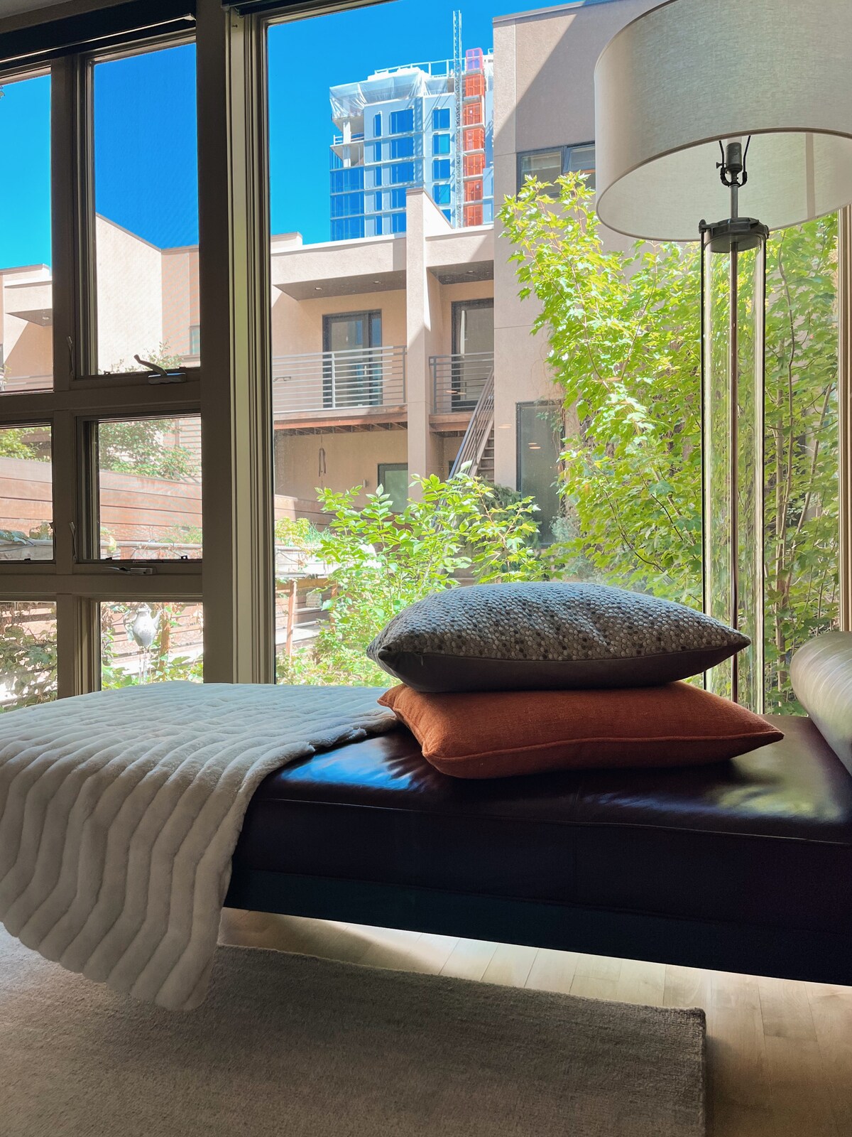 A cozy corner in the living area features a sleek chaise lounge adorned with several cushions. Natural light floods in through large windows framed by lush greenery outside, creating a serene indoor ambiance. A modern floor lamp provides additional illumination.