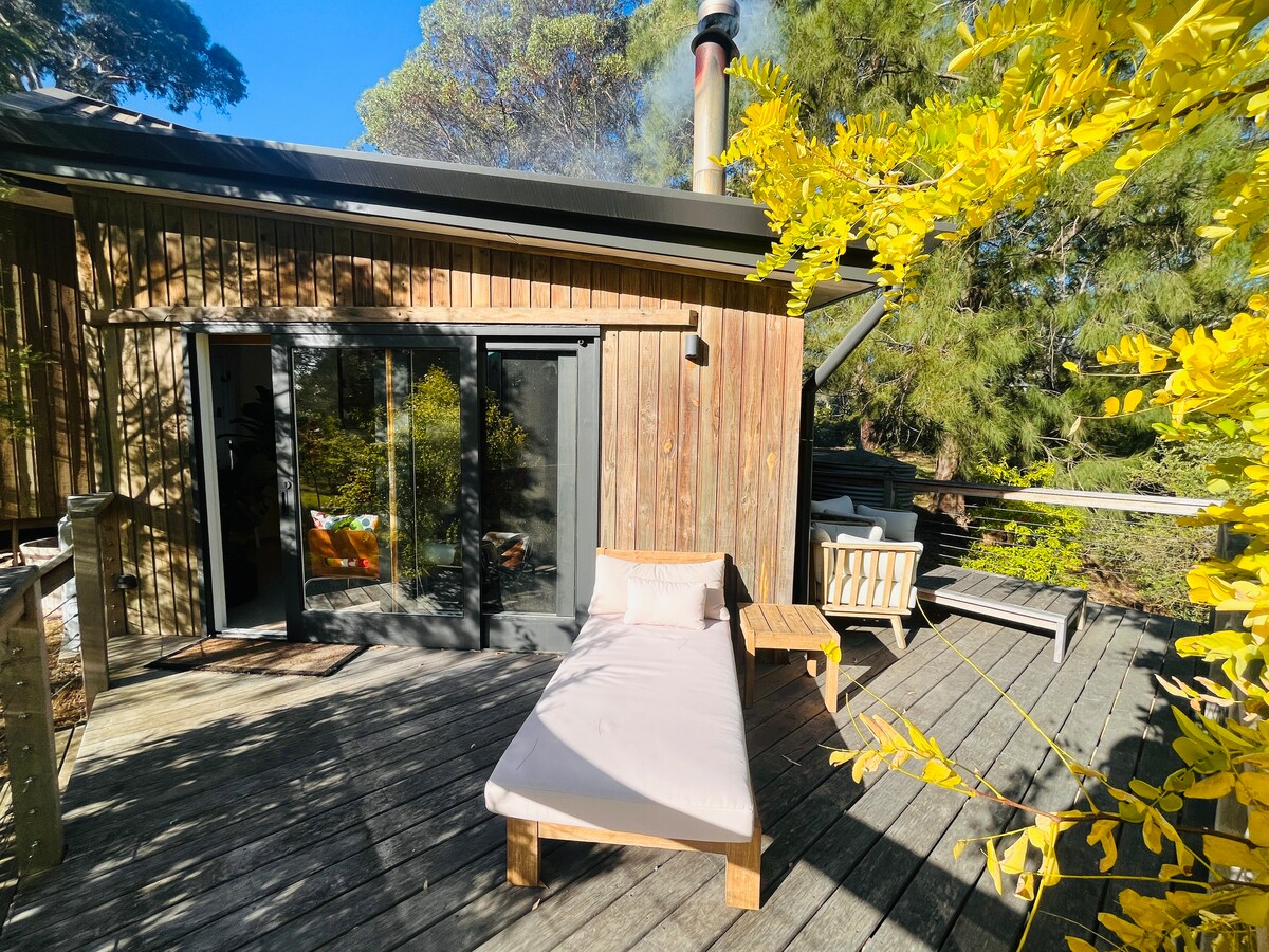 A wooden deck is surrounded by lush greenery, with a single lounge chair positioned for relaxation. Large glass doors provide access to the interior, while a small table sits nearby. The sunlight filters through the leaves, creating a serene outdoor space.