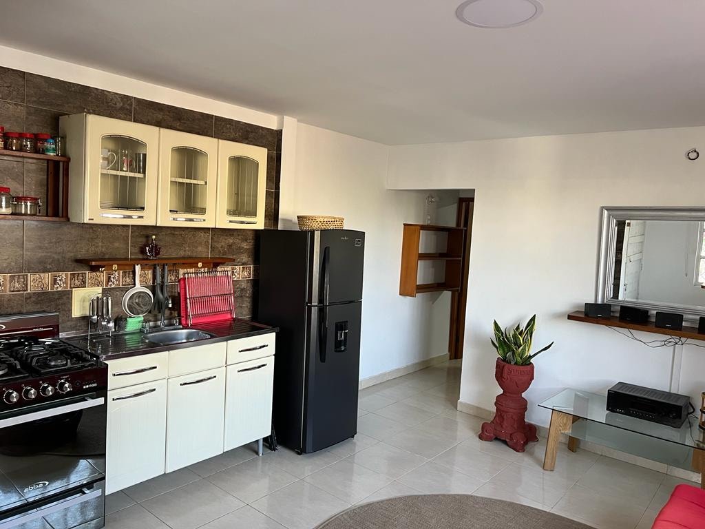A modern kitchen with white cabinetry and a dark refrigerator is featured. Cooking appliances, including a gas stove, are visible. A small breakfast table and decorative wall shelves enhance the space. Natural light enters through a nearby window, illuminating the clean, tiled floor.