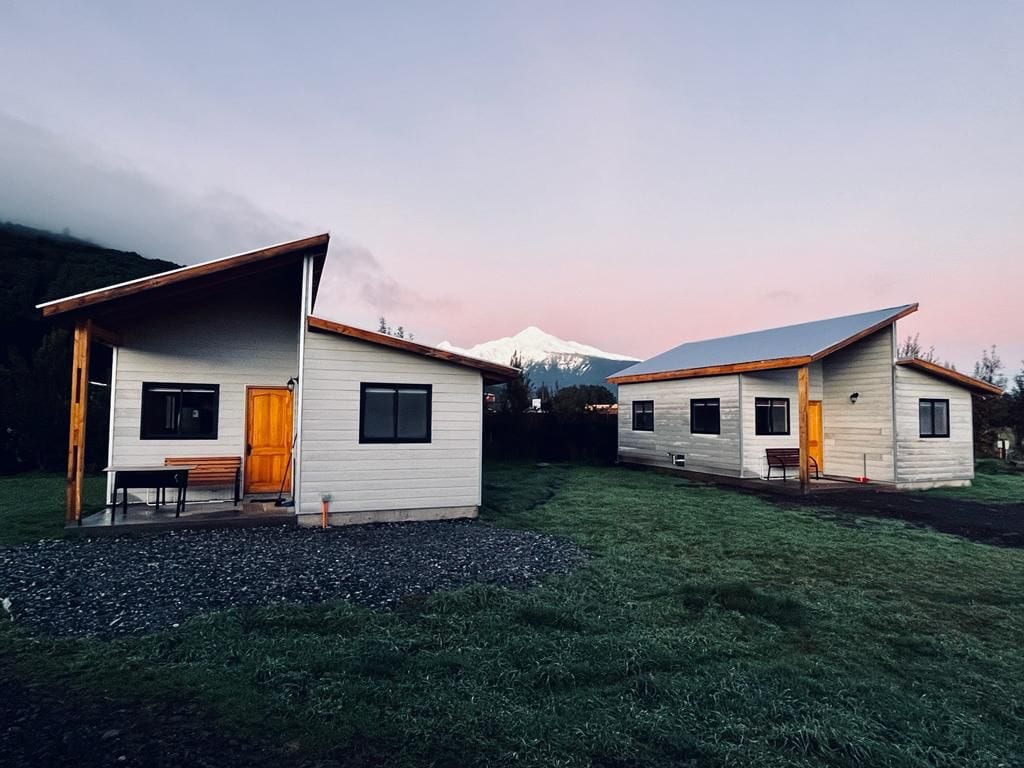 Two spacious cabins are located side by side on a grassy area. Each cabin features large windows and a front door with a bright orange finish. The snow-capped Volcán Yates is visible in the background, surrounded by soft pink hues of the dawn sky.