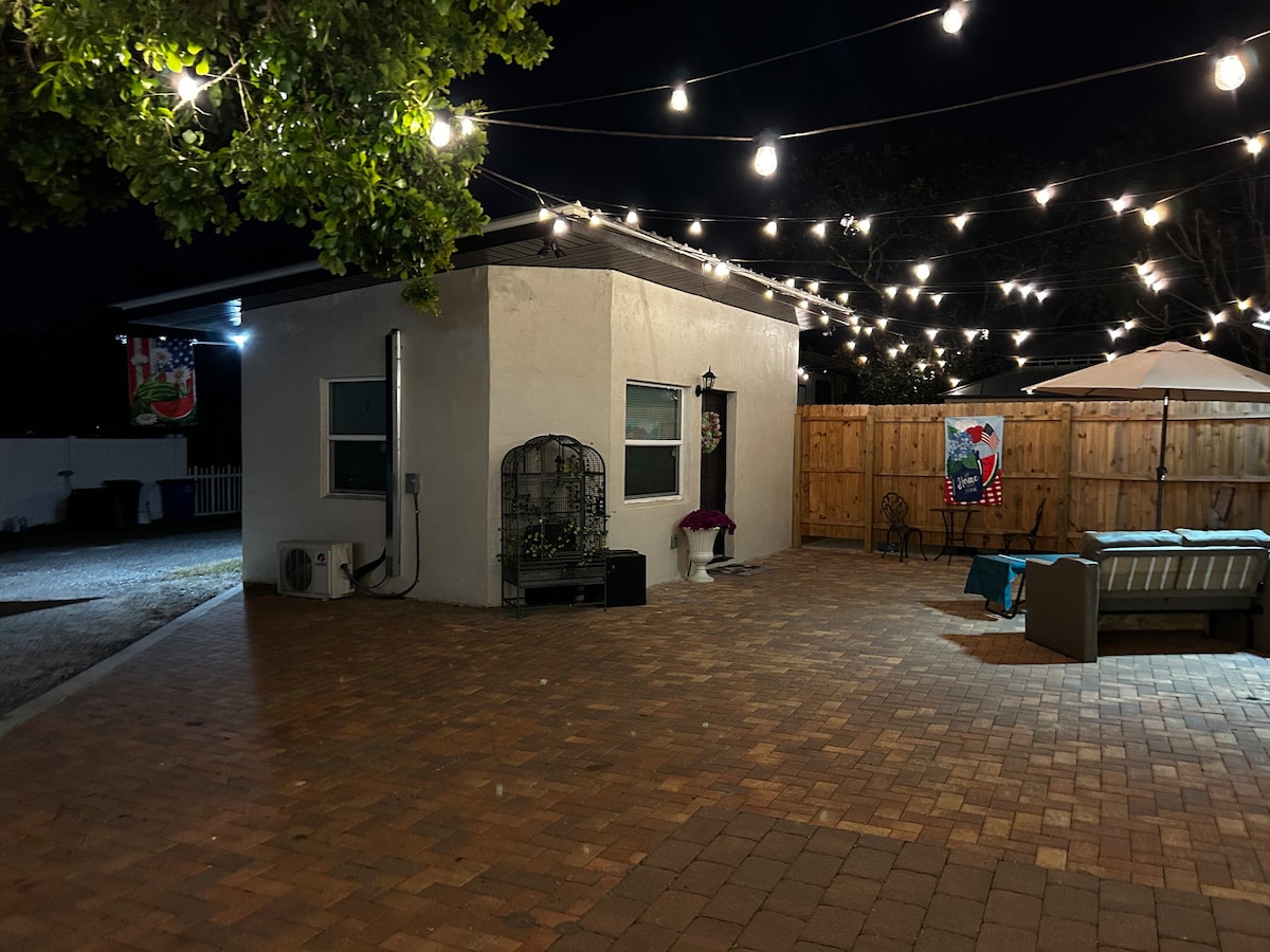 An outdoor patio area is illuminated by string lights, creating a soft glow against the evening sky. The space features a paved floor, comfortable seating arrangements, and a wooden fence that adds privacy. Decorative elements, such as wall art, enhance the inviting ambiance.