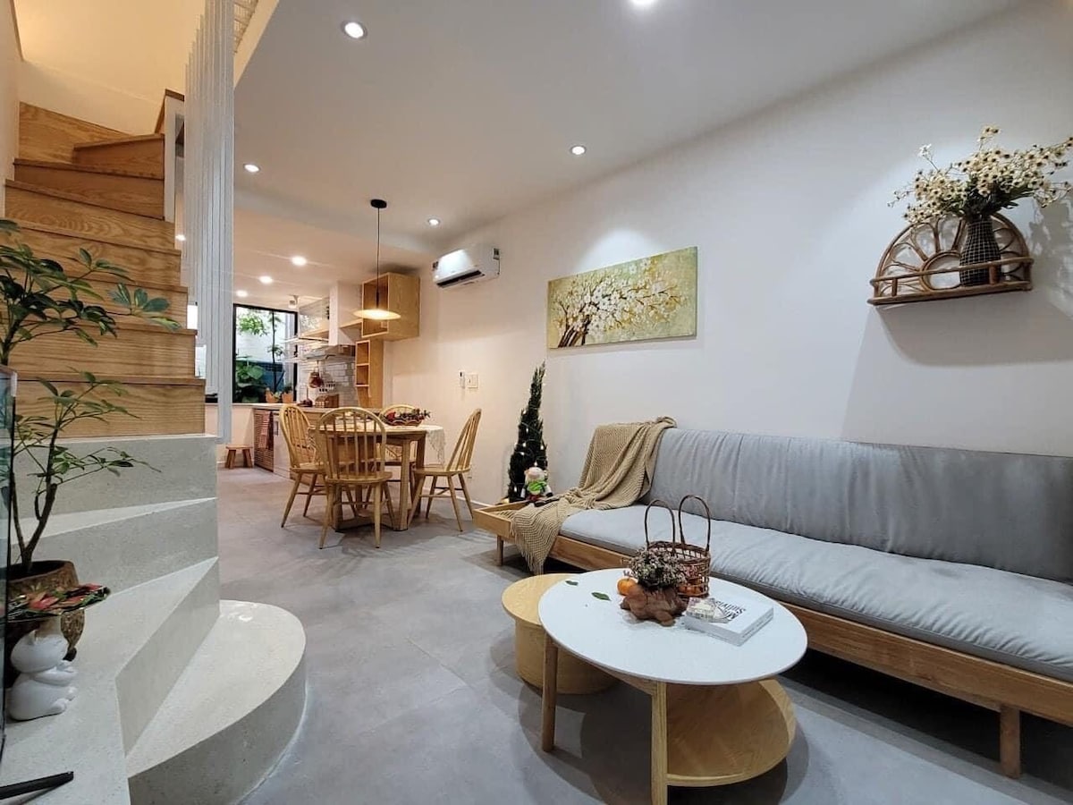 A modern living area features a light grey sofa paired with a wooden coffee table. The space is complemented by a dining table with six wooden chairs. Natural light filters through the windows, enhancing the shape of the staircase in the background.
