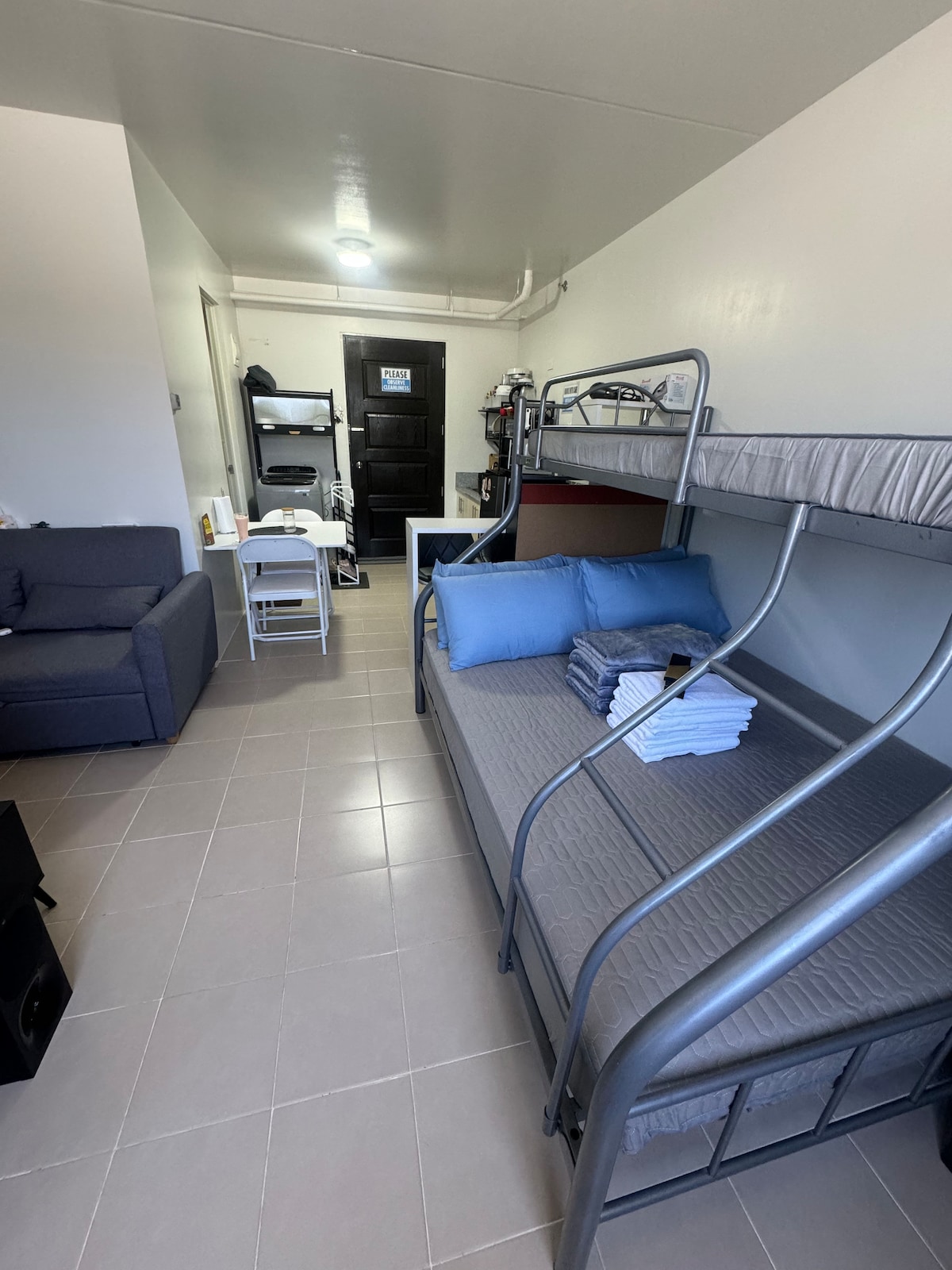 An open studio layout features a bunk bed and a single bed with neatly arranged linens. A comfortable seating area is presented with a grey sofa, and a dining table with chairs can be seen in the corner. The kitchenette is equipped with essential appliances.