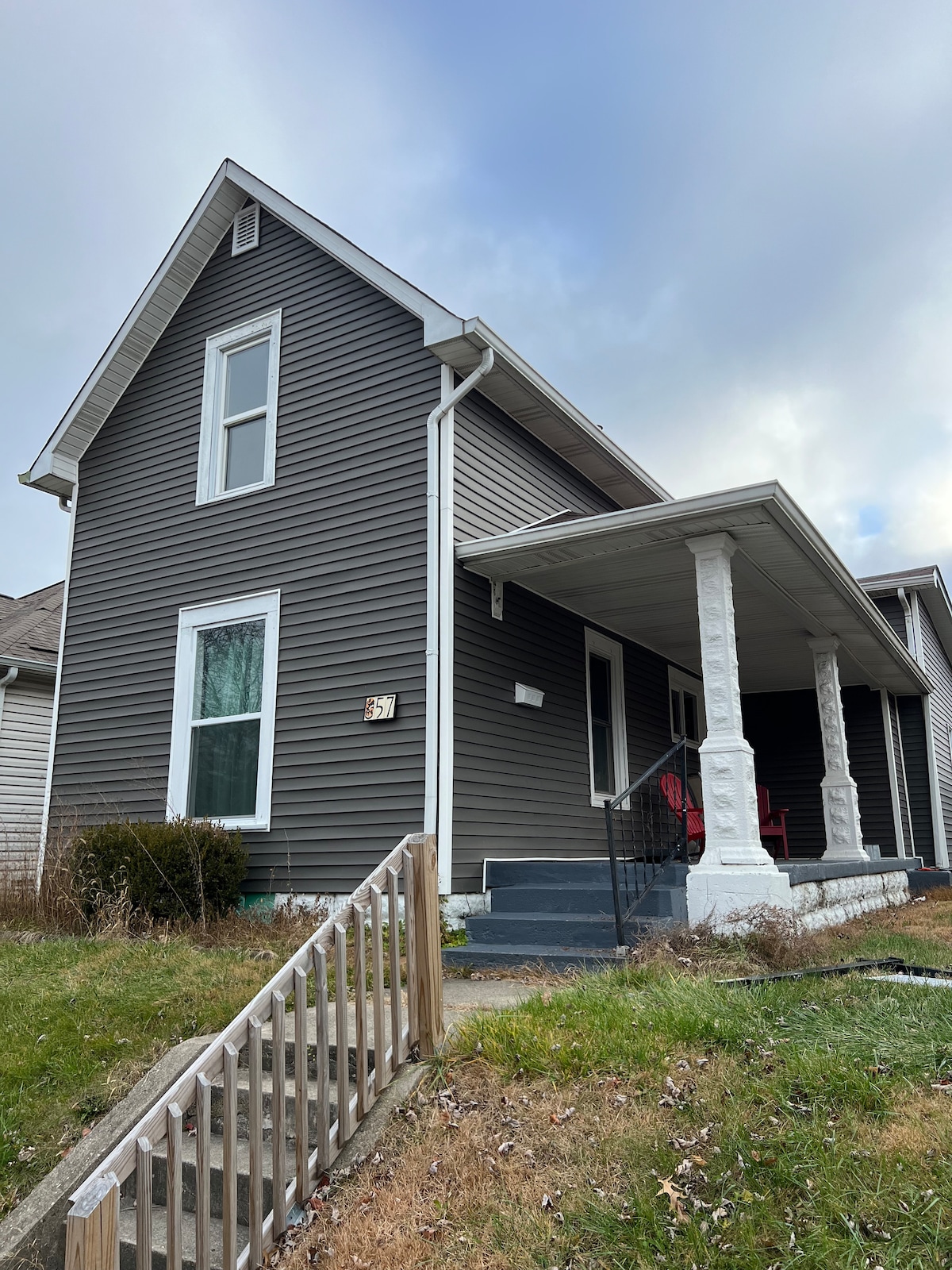 The exterior of a two-story house features gray siding and a covered porch with white columns. A set of steps leads to the entrance, while a small railing borders the pathway. Grass and shrubs are visible in the yard, under a cloudy sky.