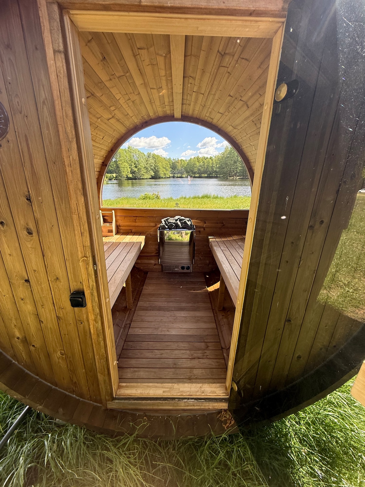 An inviting wooden sauna features an arched entryway leading to a serene view of a lake framed by lush greenery. Two long benches are positioned inside, and a small grill is visible, enhancing the outdoor experience.