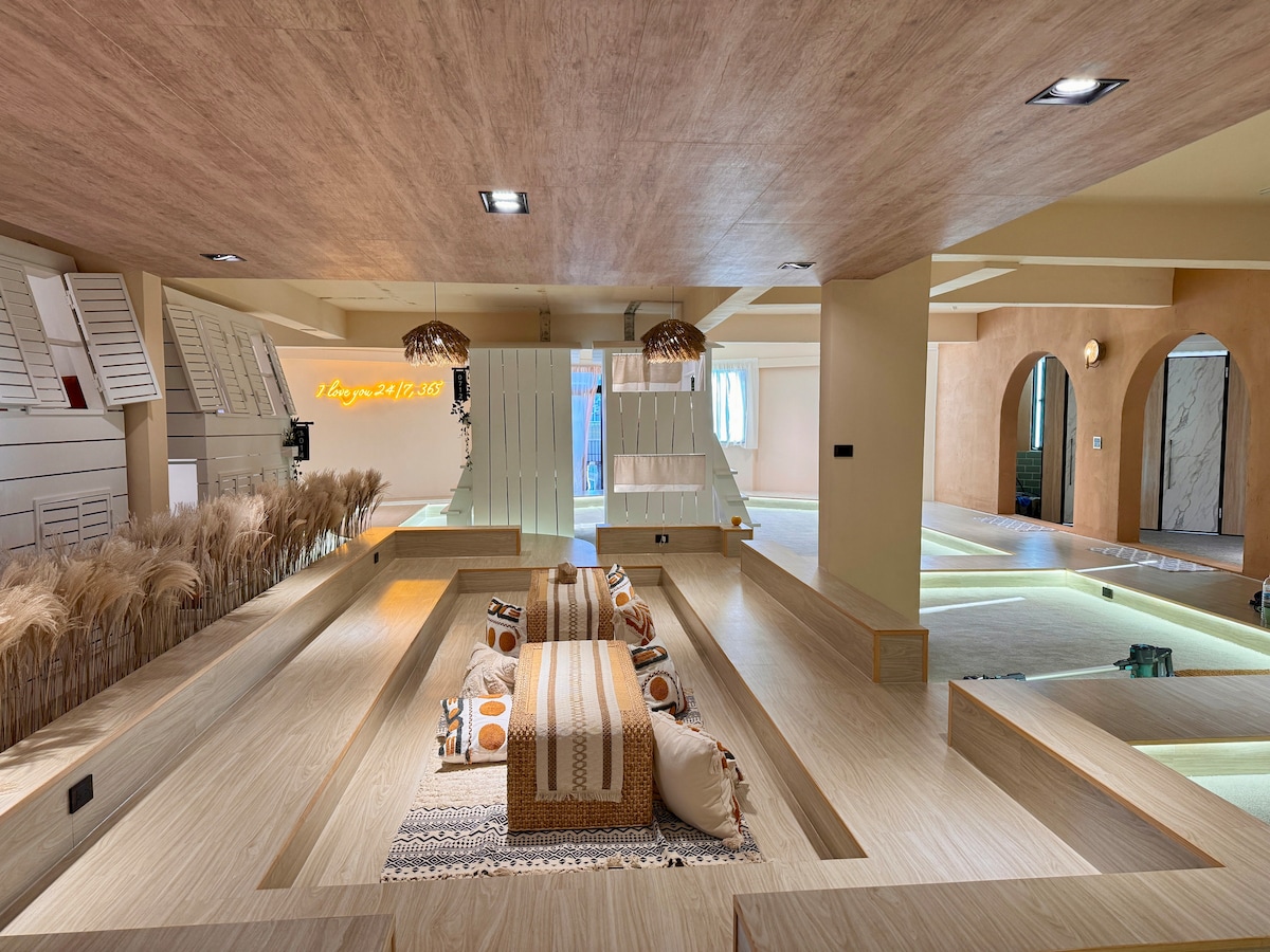 A spacious indoor area features distinct levels, accented by natural wood tones and warm lighting. Cozy seating is arranged on elevated platforms, complemented by various decorative cushions. The ambiance is complemented by softly glowing light strips and textured wall elements that add depth to the space.