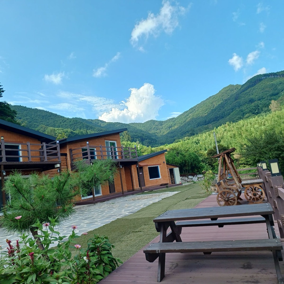 A serene outdoor view features two wooden structures set against a backdrop of lush, green mountains. A pathway made of stone leads through the garden, which is adorned with vibrant flowers. The sky is clear with a few scattered clouds, enhancing the peaceful atmosphere.