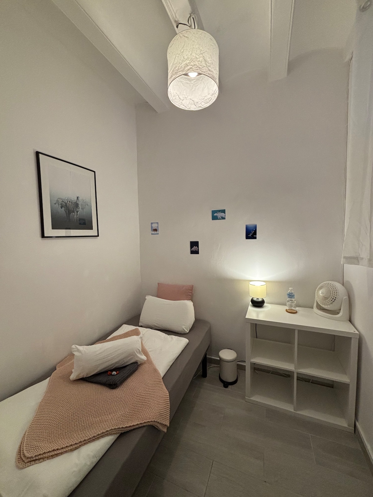 A compact room features a single bed adorned with a soft blanket and decorative pillows. A small nightstand holds a lamp and a decorative piece. The decor includes framed art on the wall and several photographs, creating a personal touch. Natural light enters through a window covered with sheer curtains.