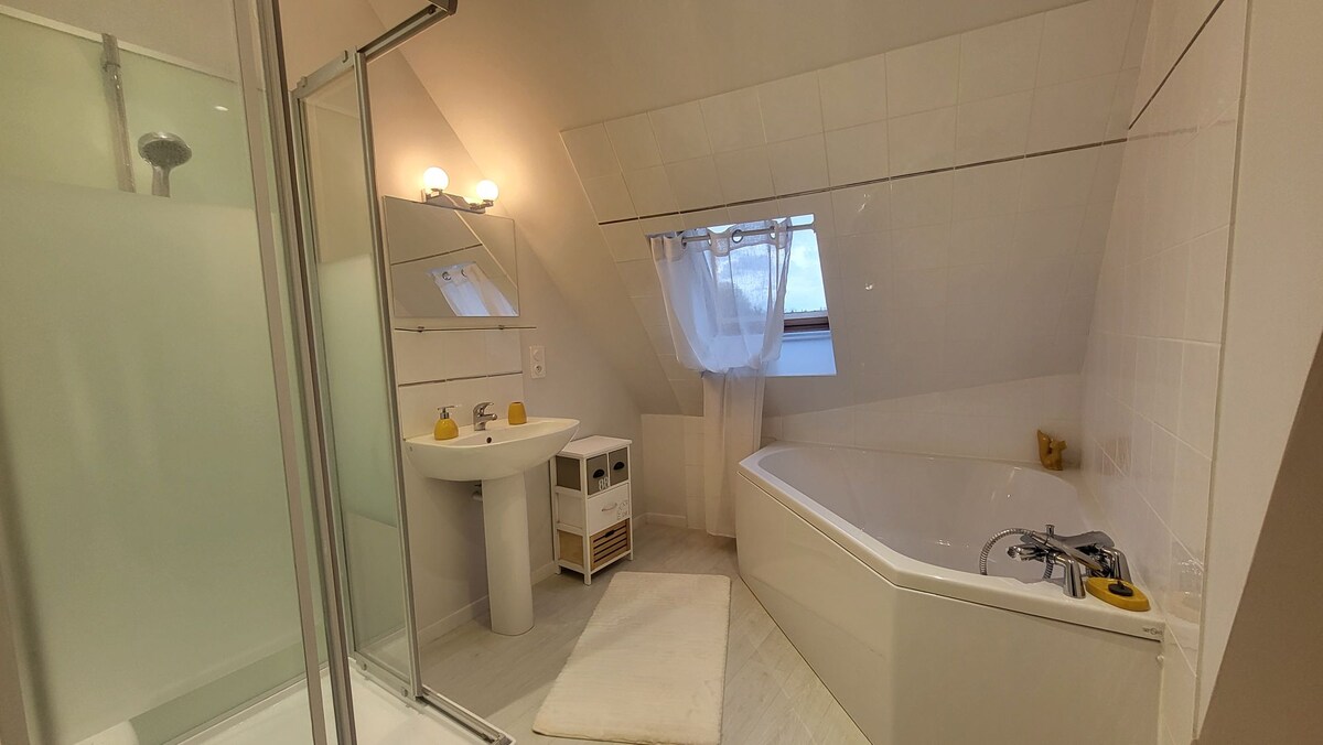 The bathroom features a spacious corner bathtub beside a large shower enclosure. A single sink with a mirror above is located next to a small storage unit. Natural light enters through a window adorned with a sheer curtain, illuminating the serene space.