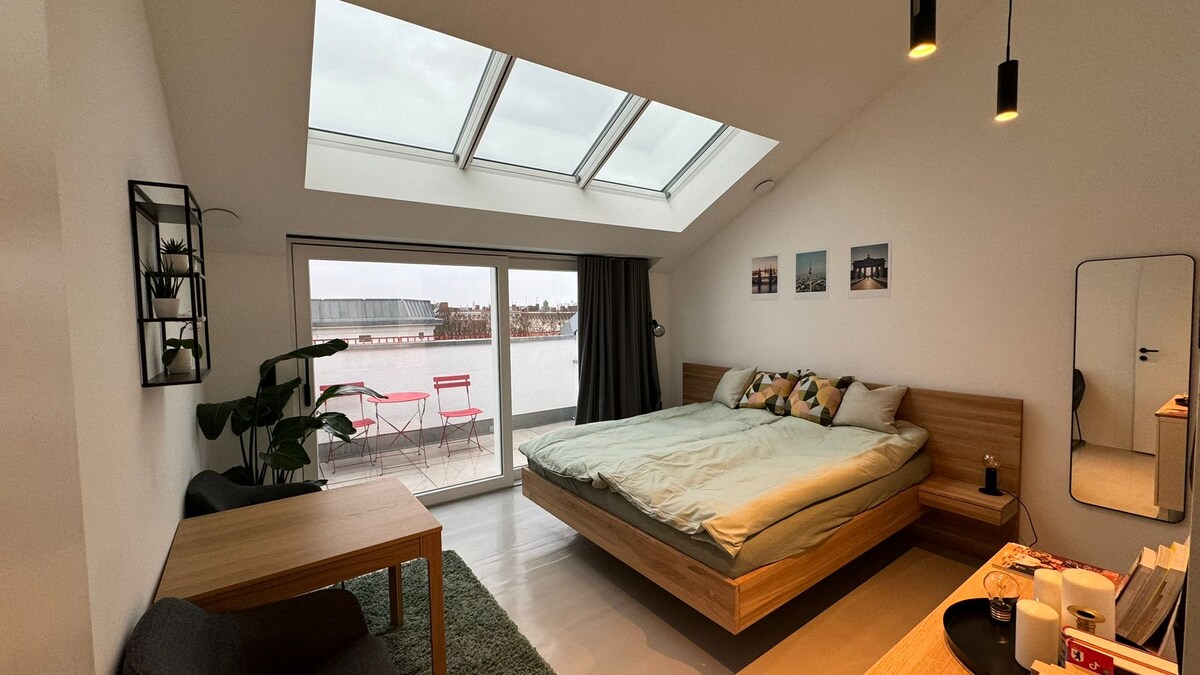 A spacious bedroom features a large bed adorned with decorative pillows, positioned under slanted skylights. Natural light fills the room, illuminating the simple wooden furnishings, including a small dining table and a mirror. A sliding door leads to a private terrace with outdoor seating.