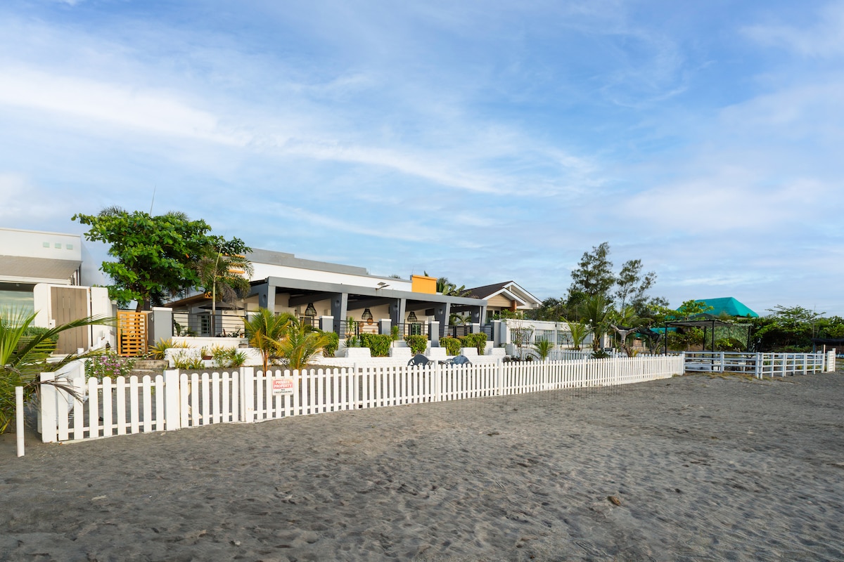 A beachfront villa is surrounded by a white picket fence, featuring a well-maintained garden with tropical plants. The structure includes two distinct sections, showcasing a modern design under a clear blue sky. The sandy beach is visible in the foreground, enhancing the coastal ambiance.
