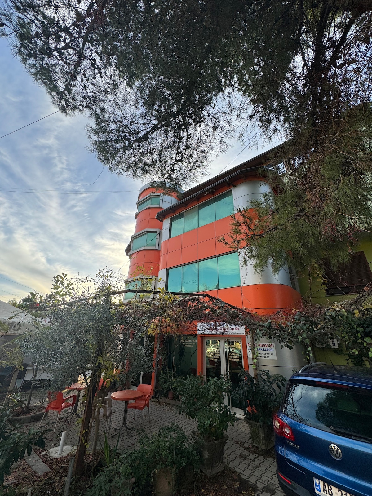 A modern multi-story building features a blend of vibrant orange and glass façades, surrounded by greenery. The ground level showcases a patio area with two round tables and chairs, inviting open-air seating under the trees.