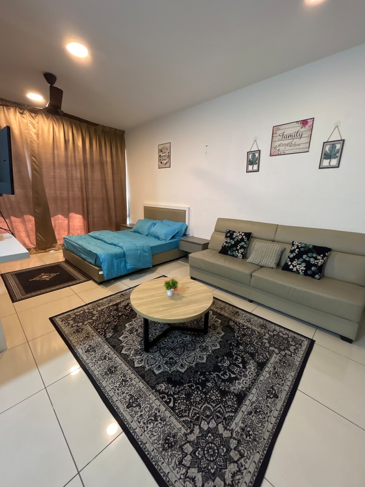 A living area features a light-colored sofa paired with patterned cushions, alongside a round wooden coffee table. A spacious bed dressed in blue linens is positioned in the background. Natural light filters through floor-to-ceiling curtains, accentuating the room's layout.