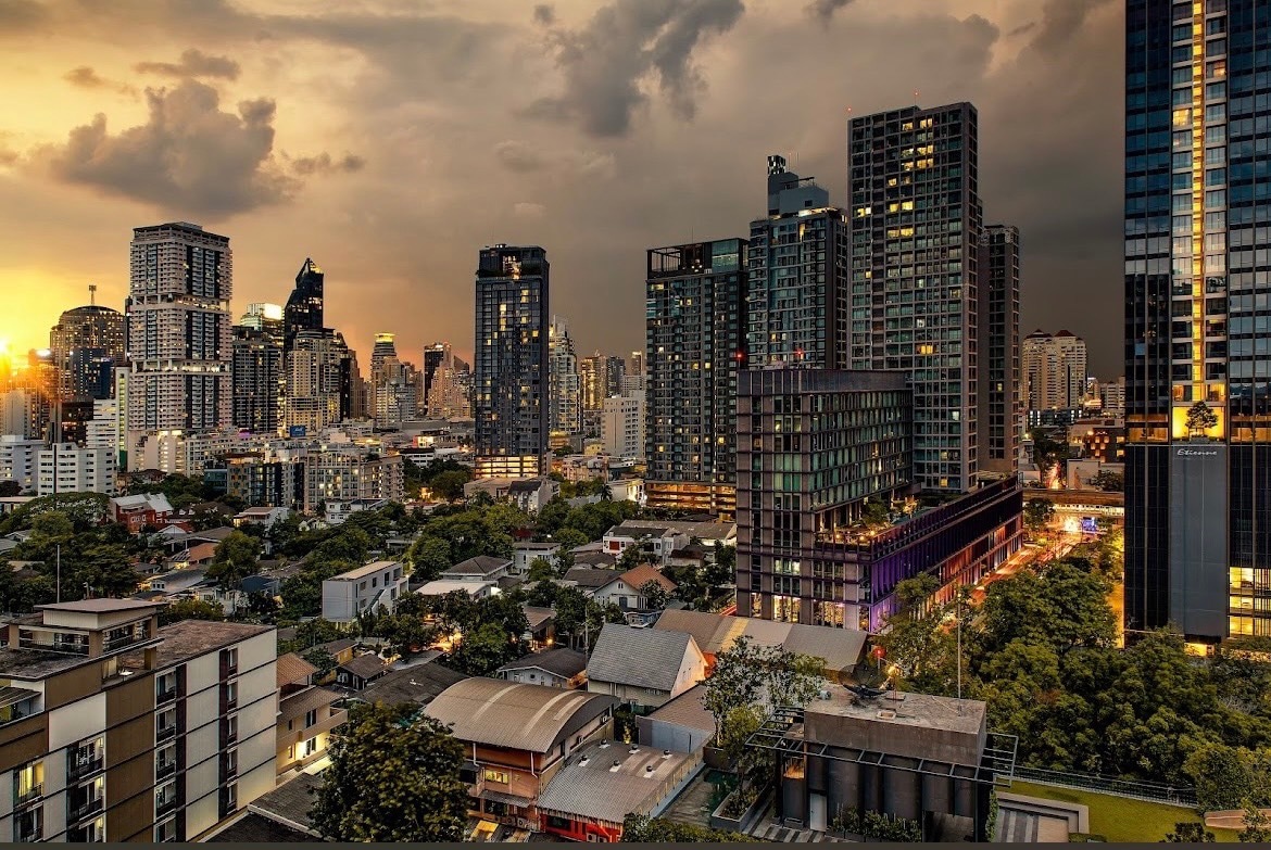 A city skyline is showcased during twilight, featuring a mix of modern high-rise buildings and lighter residential structures below. The warm glow of streetlights and reflections off windows create a serene atmosphere against a backdrop of soft clouds and distant greenery.