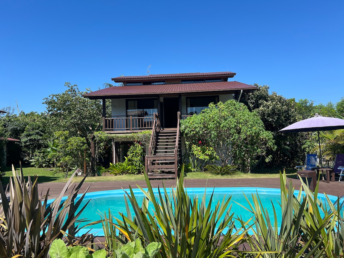 A two-story house is set amidst lush greenery, visible from a shared swimming pool. Steps lead up to a covered porch, where dark wooden railings and a gabled roof are featured. A clear blue sky complements the serene outdoor setting.