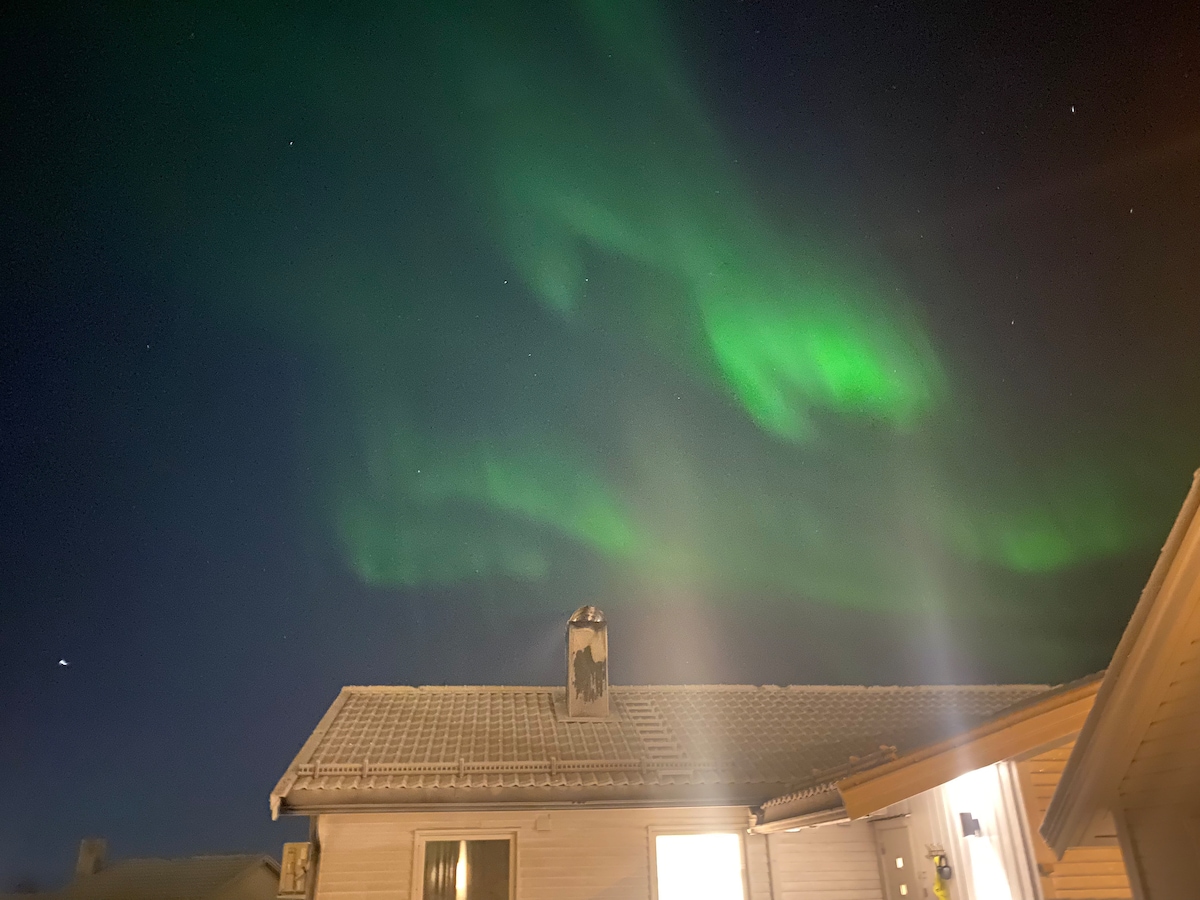 The image showcases a rooftop under a clear night sky, featuring vibrant green auroras dancing above. A chimney is visible, adding to the cozy atmosphere of the home. Stars can be seen scattered throughout the deep blue sky.
