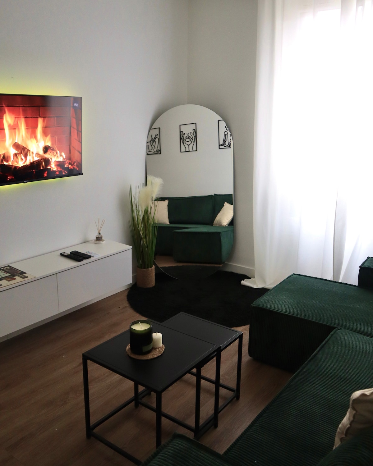 A cozy living area features a green sofa and armchair arranged around a black coffee table. A wall-mounted television displays a roaring fire, adding warmth to the space. Natural light filters through sheer curtains, illuminating decorative plants and minimalistic artwork.