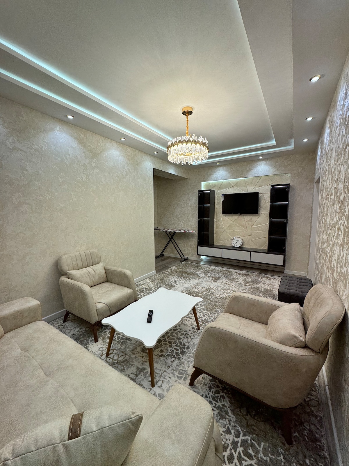A modern living area is showcased, featuring a plush seating arrangement with two beige armchairs and a beige sofa. A sleek, light-colored coffee table sits in the center. A chandelier hangs from the ceiling, and a wall-mounted television is present, accompanied by a decorative shelving unit.