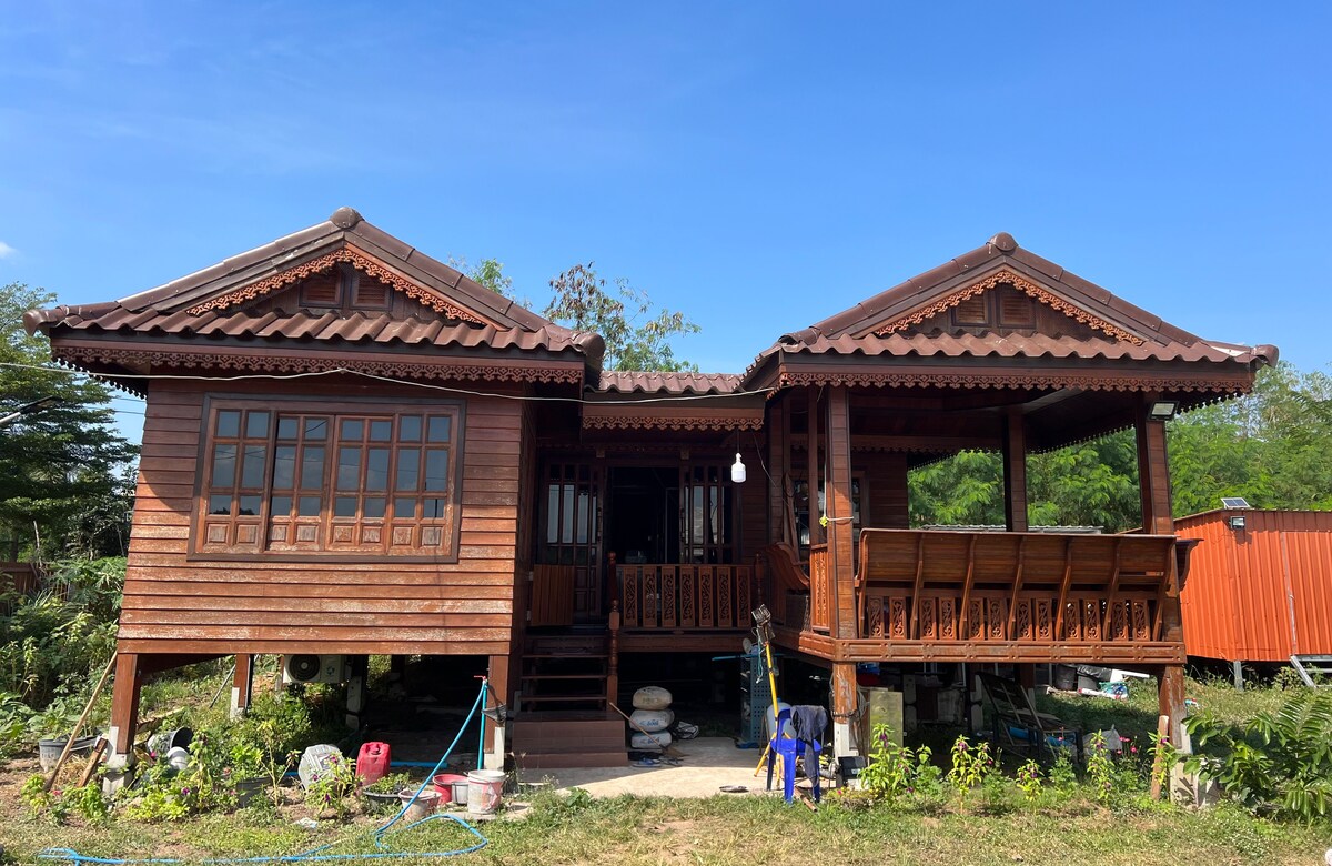 A traditional wooden house with slanted roofs and intricate carvings is showcased against a clear blue sky. The structure features a welcoming porch with wooden railings, surrounded by lush greenery and outdoor furniture placed on the ground below.