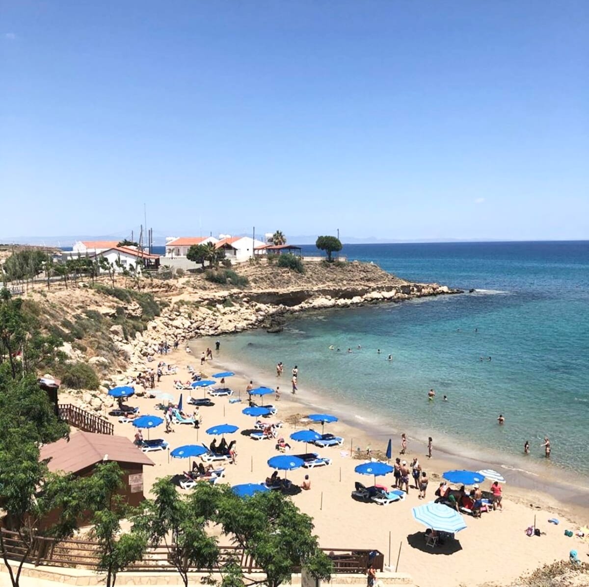 A sandy beach is visible, dotted with blue umbrellas providing shade for sunbathers. The clear water is seen calmly lapping at the shore, while small rocky cliffs frame the beach. Beachgoers can be spotted enjoying the sun and swimming in the ocean.