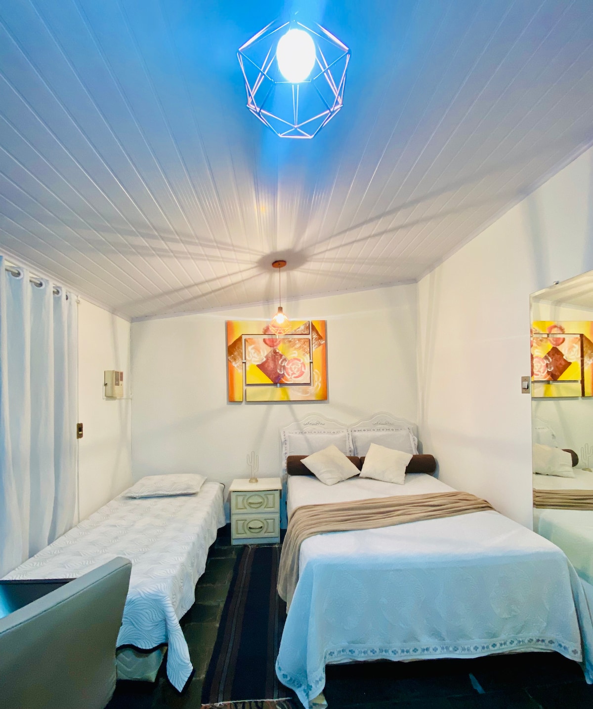 A well-lit sleeping area features a spacious double bed alongside a single bed, both dressed in white bedding. Above, a modern light fixture casts a soft glow. Decorative artwork is displayed on the wall, and a mirror reflects the room's spaciousness.