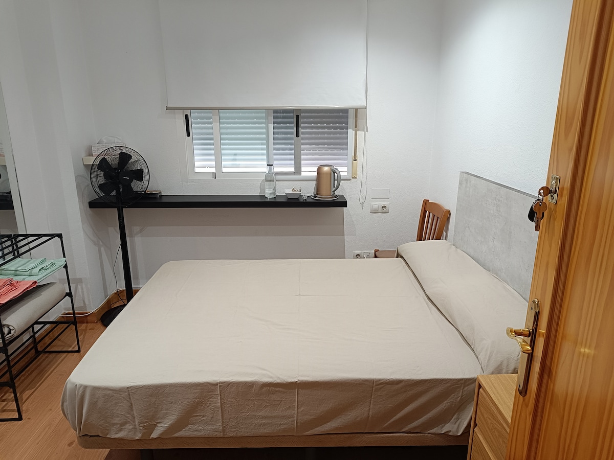 A simple sleeping area features a spacious bed with a neutral-colored bedspread. A fan is positioned nearby, and a small table with a kettle is visible against the wall. Natural light filters in through a window covered by light-colored blinds.