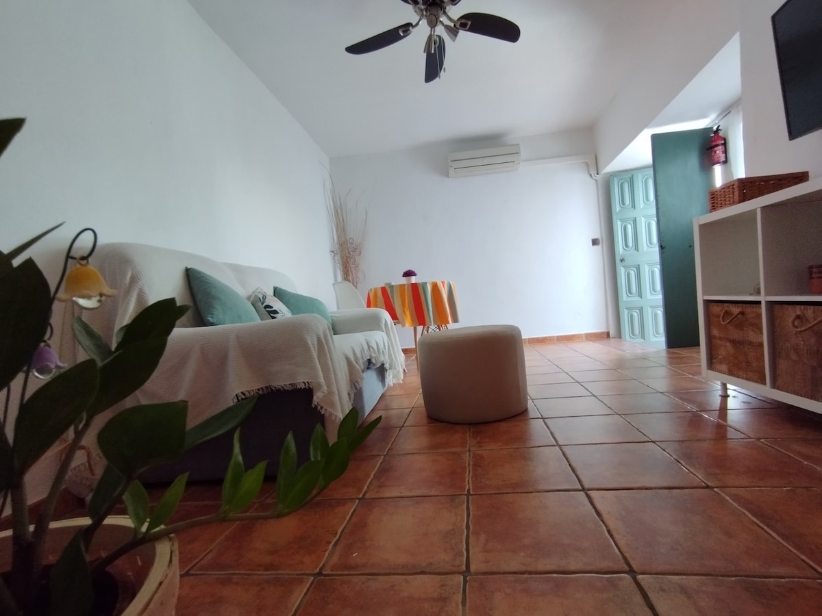 A bright living area is presented, featuring a comfortable chaise lounge and a round dining table adorned with colorful tableware. The tiled floor reflects warm tones, while a ceiling fan is visible above, contributing to a relaxing atmosphere. Light enters through an adjacent door, highlighting the space.