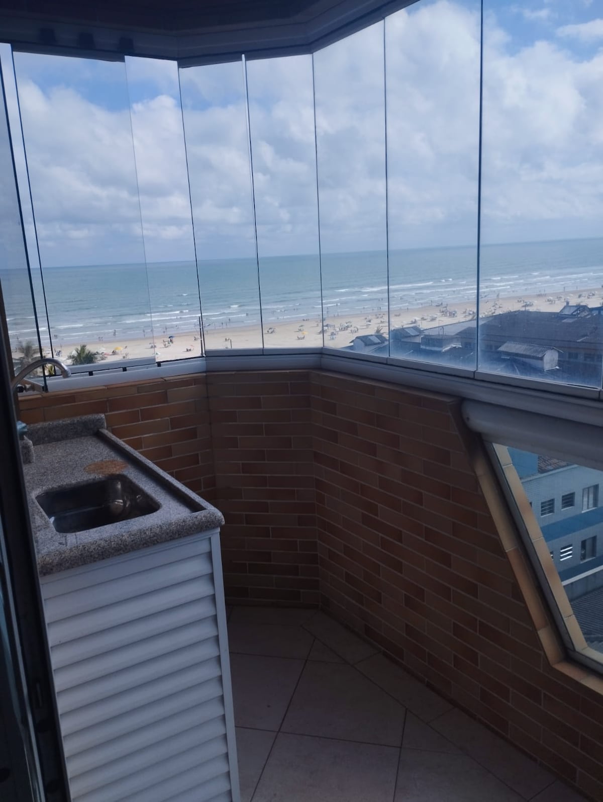 A balcony offers a clear glass enclosure, providing an unobstructed view of the ocean and nearby beach. A small kitchen area with a granite countertop and sink is visible, creating a functional space for outdoor cooking and entertaining.