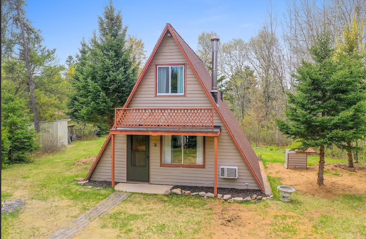 A charming A-frame cabin is presented with a wooden front porch and an angled roof. Large windows allow natural light, and a cozy entrance leads to the green grass surrounding the property. Twin evergreen trees frame the space, enhancing the serene outdoor setting.