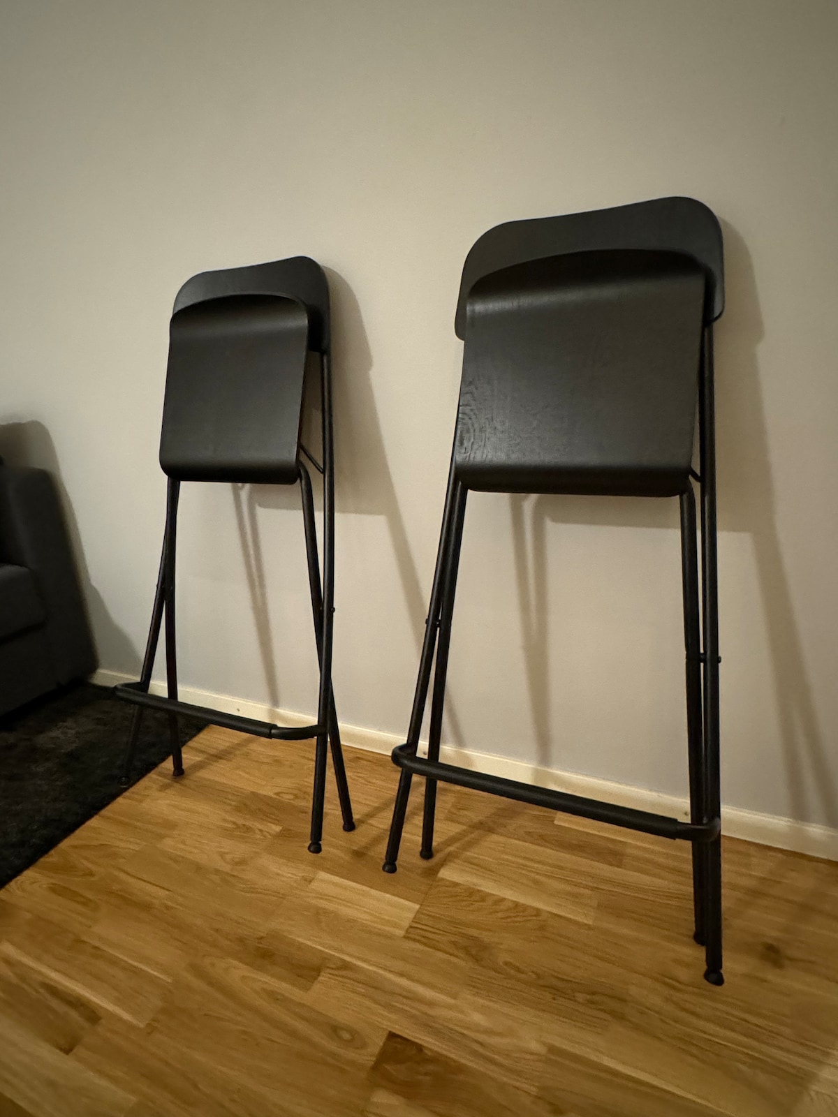 Two black, modern folding chairs are positioned against a neutral wall. They stand on a wooden floor, showcasing a minimalist design with sleek lines and a simple structure.
