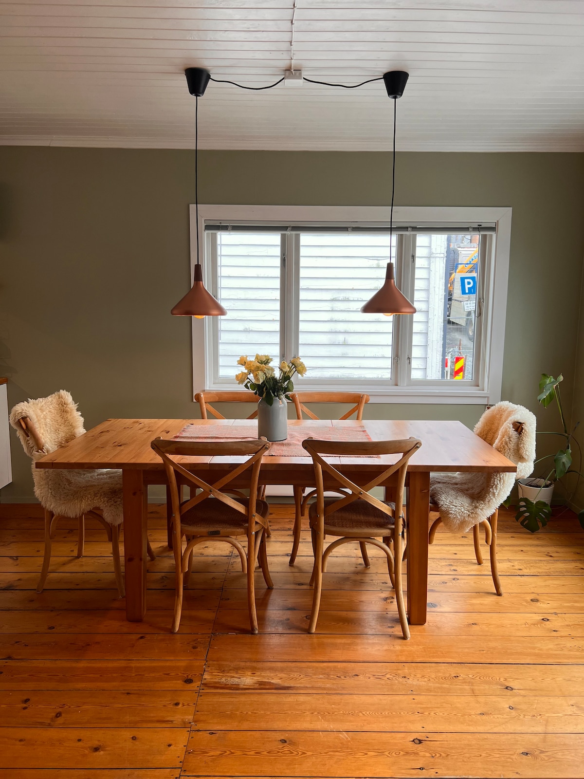 A dining area features a wooden table surrounded by several chairs. Two stylish pendant lights hang above the table, and a vase of flowers is centered on the table. Natural light enters through a large window, highlighting the warm wooden floor.