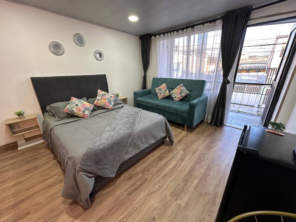 A cozy sleeping area features a spacious bed with soft gray bedding and decorative pillows. A teal sofa adds a pop of color, positioned near large windows dressed in light curtains. Wooden flooring enhances the inviting appearance of the room.