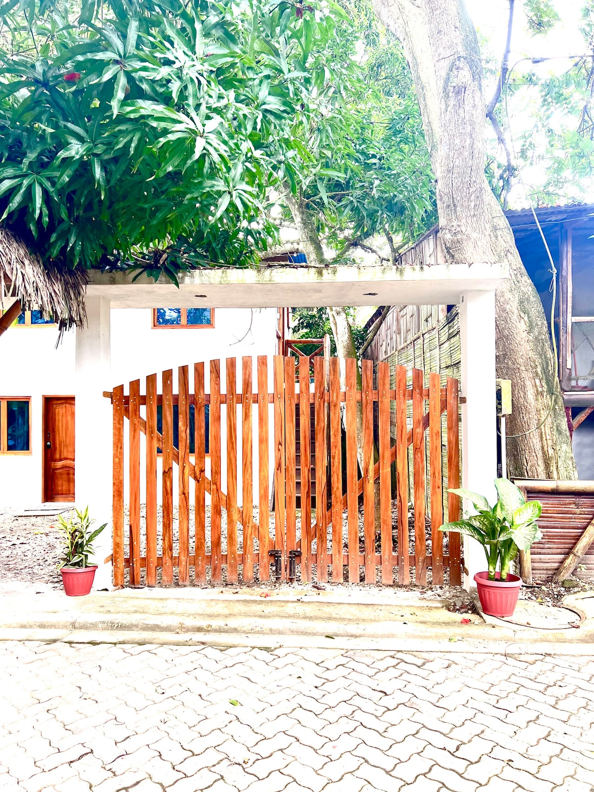 A wooden gate, designed with horizontal slats, serves as an entrance to the property. Potted plants are positioned on either side of the gate, and lush greenery surrounds the area, providing a sense of privacy and nature.