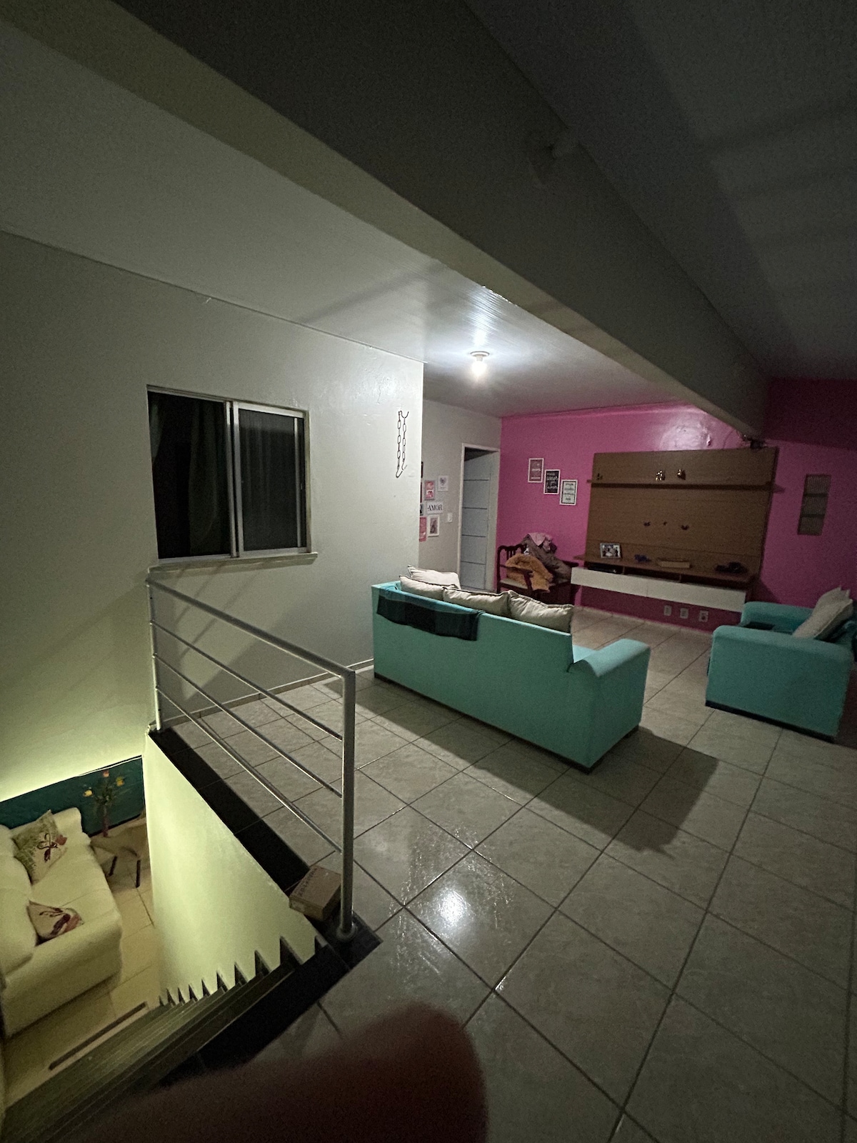 An open living space features light-colored walls with one accent wall in pink. Two turquoise sofas are positioned adjacent to a sleek entertainment unit. Natural light enters through a nearby window, illuminating the polished tile floor. A staircase leads to an upper level.