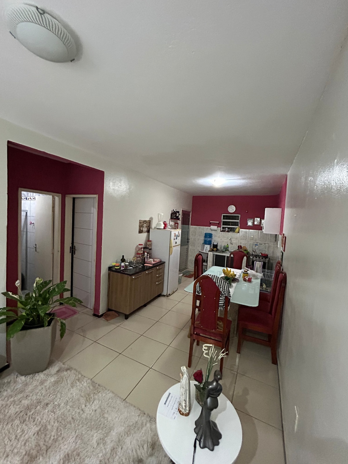 An open layout reveals a living area with a white rug and a small dining table set for six. A kitchen with a refrigerator and a gas stove is visible in the background. Decorative plants add a touch of greenery to the space, enhancing its inviting appearance.