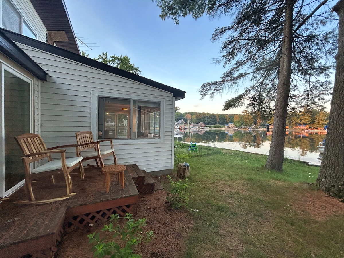 A small outdoor deck features two wooden chairs, positioned to overlook the serene lake. Pine trees frame the scene, which showcases calm water reflecting the surrounding landscape. The attached home has windows that provide expansive views of the lakeside setting.