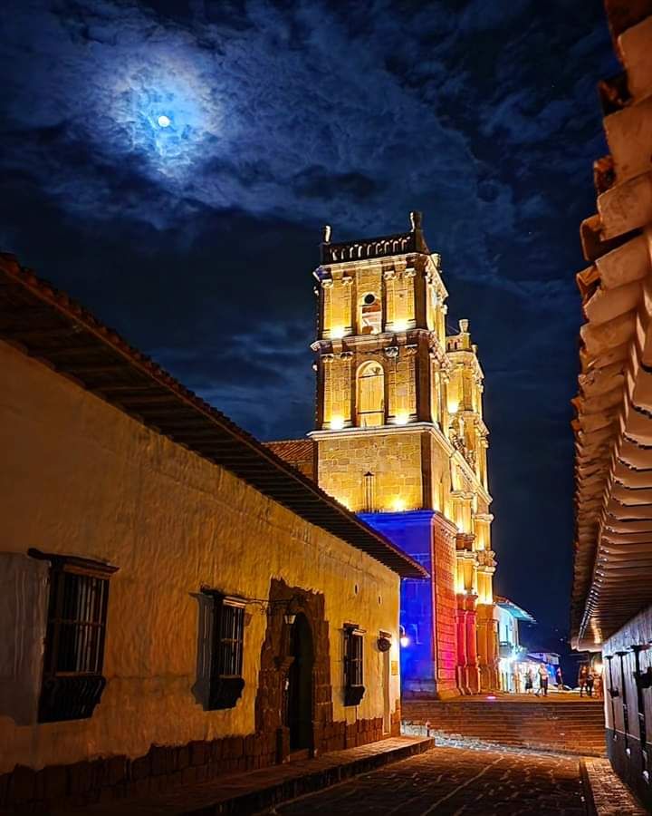 A historic tower is illuminated against a night sky, casting a warm glow onto the surrounding cobblestone street. The building features artistic lighting, enhancing the texture of the stone facade. A full moon adds a serene atmosphere to the scene.