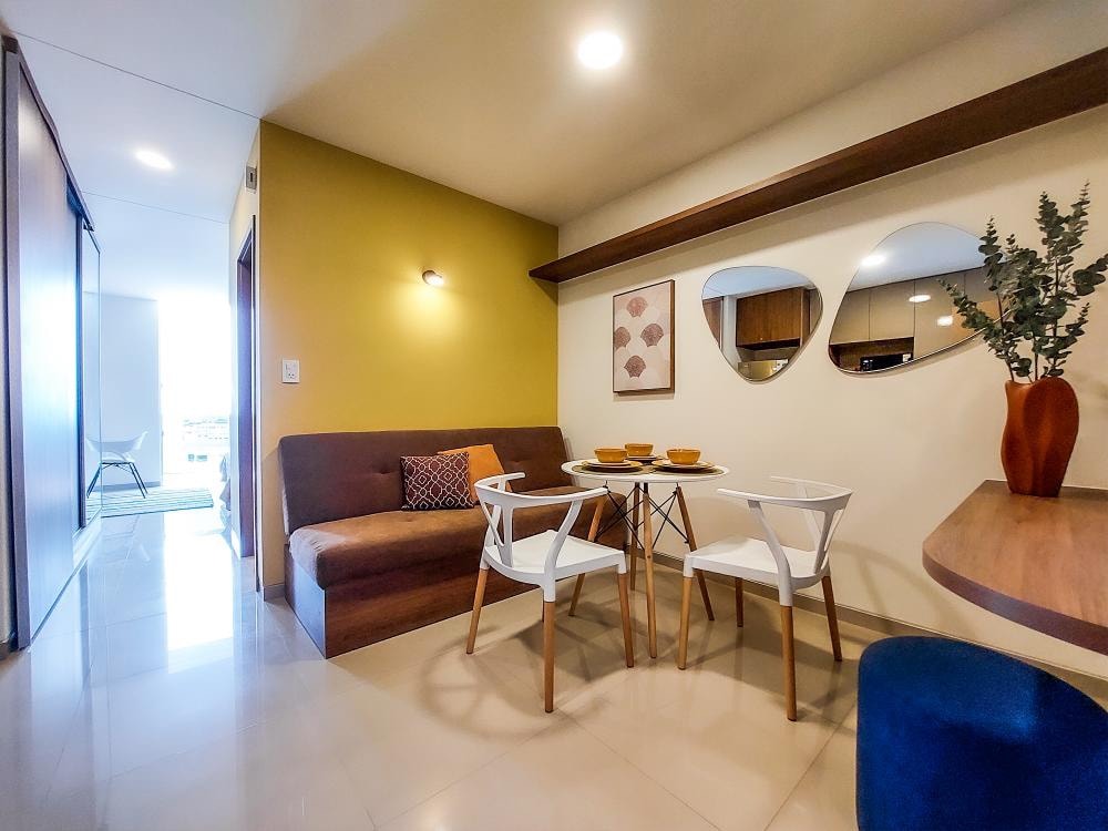 A welcoming dining area is displayed, featuring a simple white table surrounded by four chairs. A cozy seating nook with cushions is situated against a textured wall. Decorative mirrors and a vase with greenery add subtle accents to the space, enhanced by soft overhead lighting.