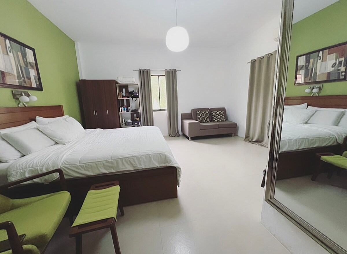 A spacious bedroom features a large bed with crisp white linens, complemented by a comfortable seating area with a sofa. Green accent walls add a touch of color, while a large mirror reflects the natural light streaming in from the window, creating a bright atmosphere.